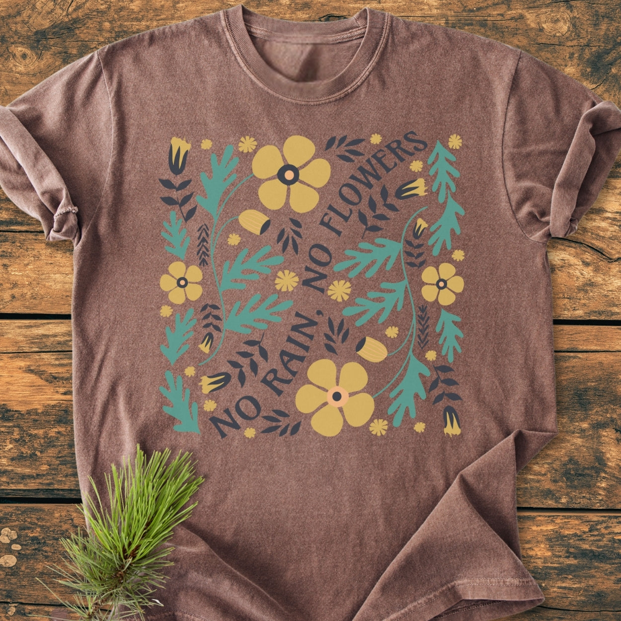No Rain No Flowers - Vintage Wash Tee features a stylized floral graphic with yellow blossoms and teal foliage, printed on a brown T-shirt, laid on a rustic wooden surface with a pine sprig accent.