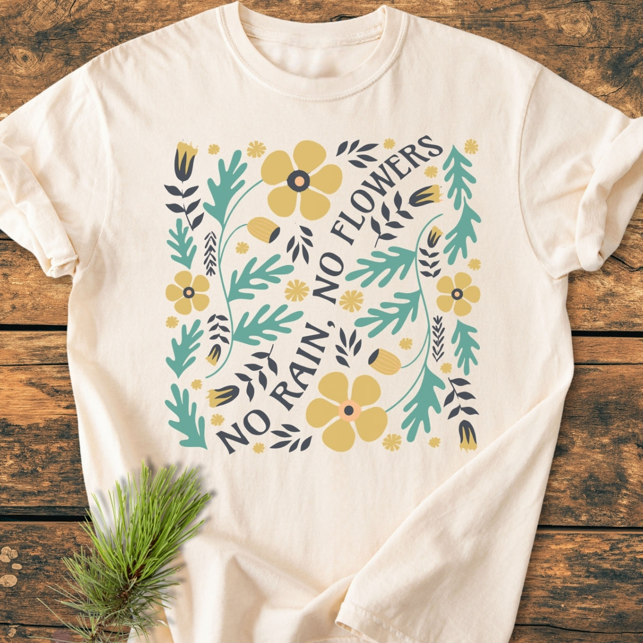 No Rain No Flowers - Vintage Wash Tee displayed on wood, featuring botanical artwork with teal leaves and mustard-yellow flowers, emphasizing the slogan. A small pine sprig adds a rustic touch.