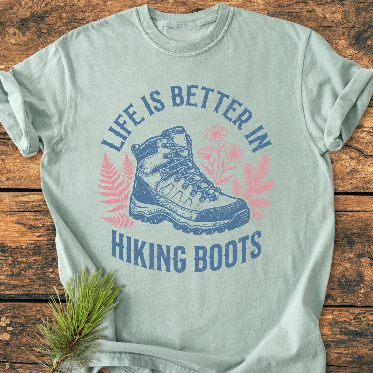 Life Is Better - Vintage Wash Tee featuring a hiking boot illustration with fern and wildflower graphics, laid on wood, enhanced by a pine sprig. Cozy, relaxed fit, 100% ring-spun cotton.