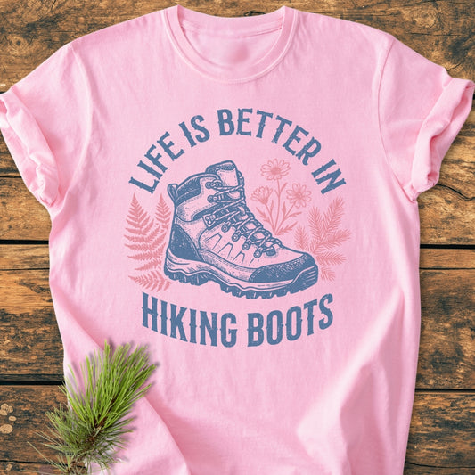 Life Is Better - Vintage Wash Tee displayed on wood, features a hiking boot print with ferns and wildflowers, and the slogan 'LIFE IS BETTER IN HIKING BOOTS,' complemented by a pine needle sprig.
