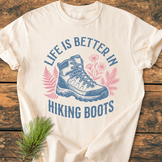 Life Is Better - Vintage Wash Tee featuring a stylized blue hiking boot with pink fern and flower accents, displayed on wood with a pine sprig. Made from 100% ring-spun cotton for comfort.