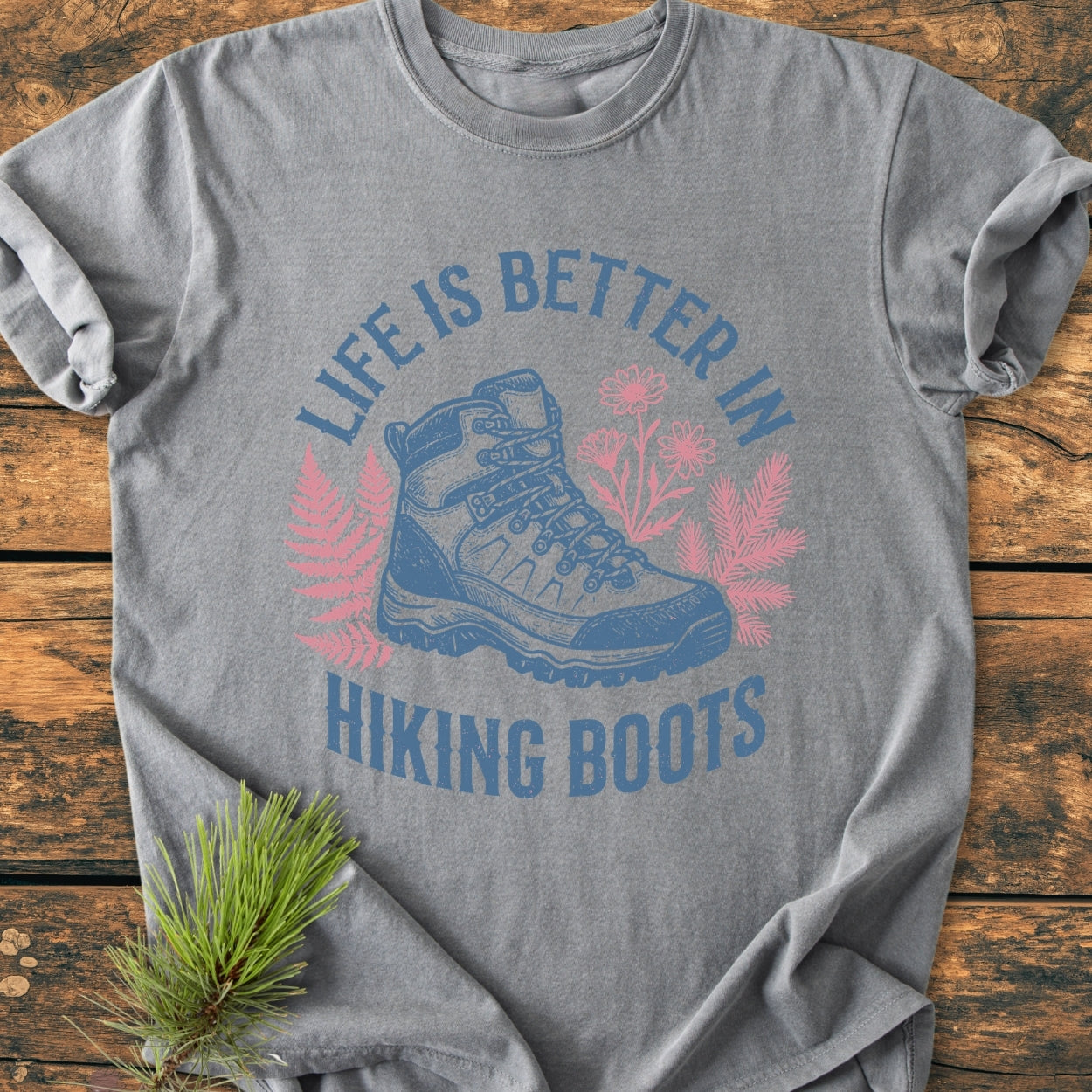 Life Is Better - Vintage Wash Tee features a hiking boot graphic with ferns and wildflowers on a gray shirt, emphasizing outdoor adventure. Laid flat with a pine branch accent, showcasing its relaxed, cozy style.