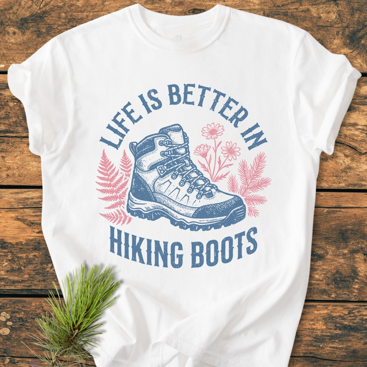 Life Is Better - Vintage Wash Tee features a hand-drawn hiking boot illustration with foliage and slogan, laid on wood surface, styled with a pine sprig. Made from soft, durable, garment-dyed cotton.