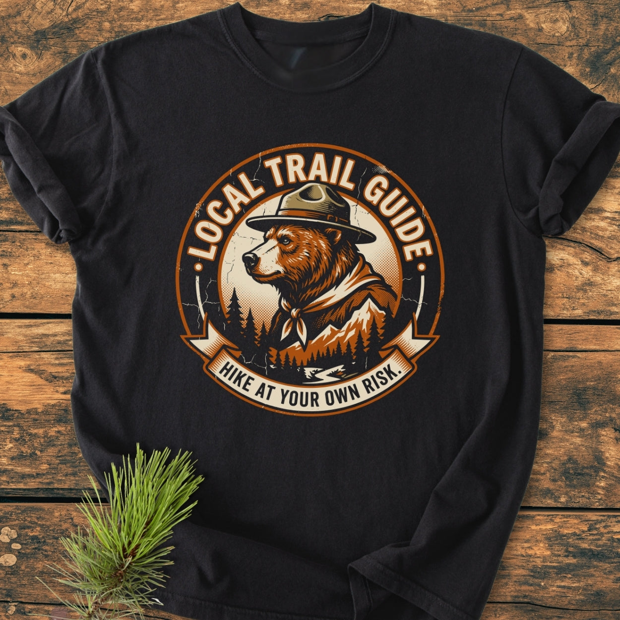 Local Trail Guide - Vintage Wash Tee featuring a circular logo with a bear in a ranger hat; laid on wood with pine, showcasing its relaxed fit and cozy, garment-dyed fabric.