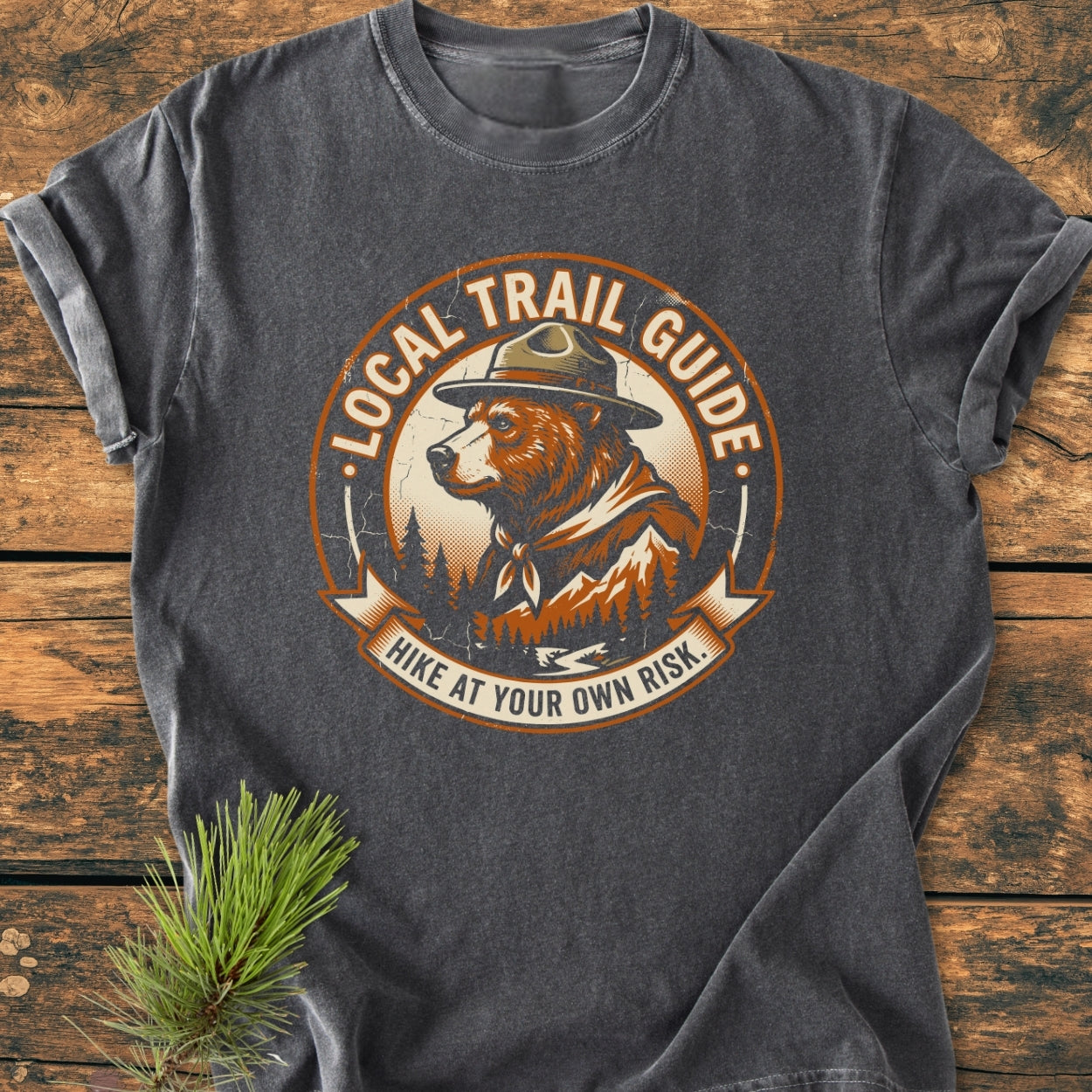 Local Trail Guide - Vintage Wash Tee featuring a bear graphic with a ranger hat, pine trees, and mountain silhouette, laid on wood. The soft, relaxed-fit fabric ensures comfort and durability.