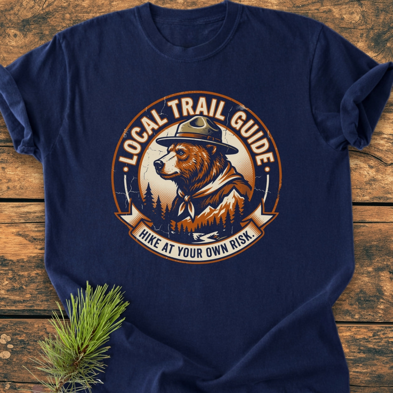 Local Trail Guide - Vintage Wash Tee featuring a circular logo with a bear in a ranger’s hat against pine trees and mountains, laid on wood; offers a relaxed fit and durable, soft fabric.