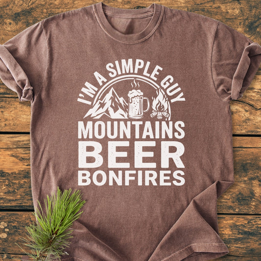 Mountains Beer Bonfires - Vintage Wash Tee featuring white text and icons of mountains, a beer mug, and campfire on a brown shirt, styled with a pine sprig on a wooden surface.