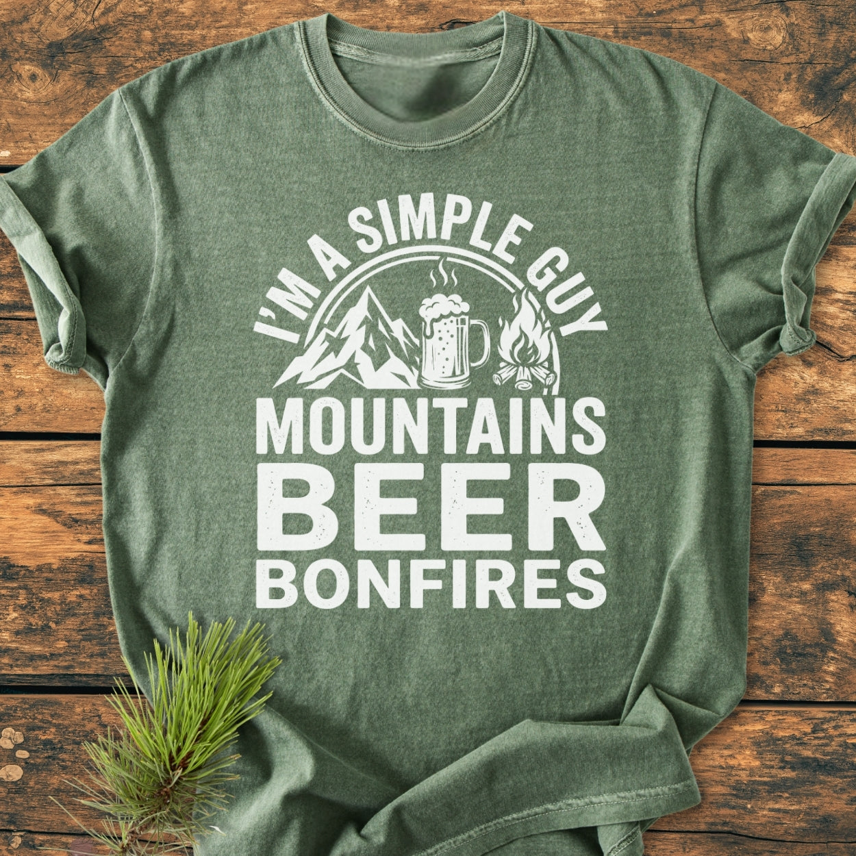 Mountains Beer Bonfires - Vintage Wash Tee, heather-green, with white slogan and icons of mountains, beer mug, and campfire. Laid on wood, features a pine sprig accent.