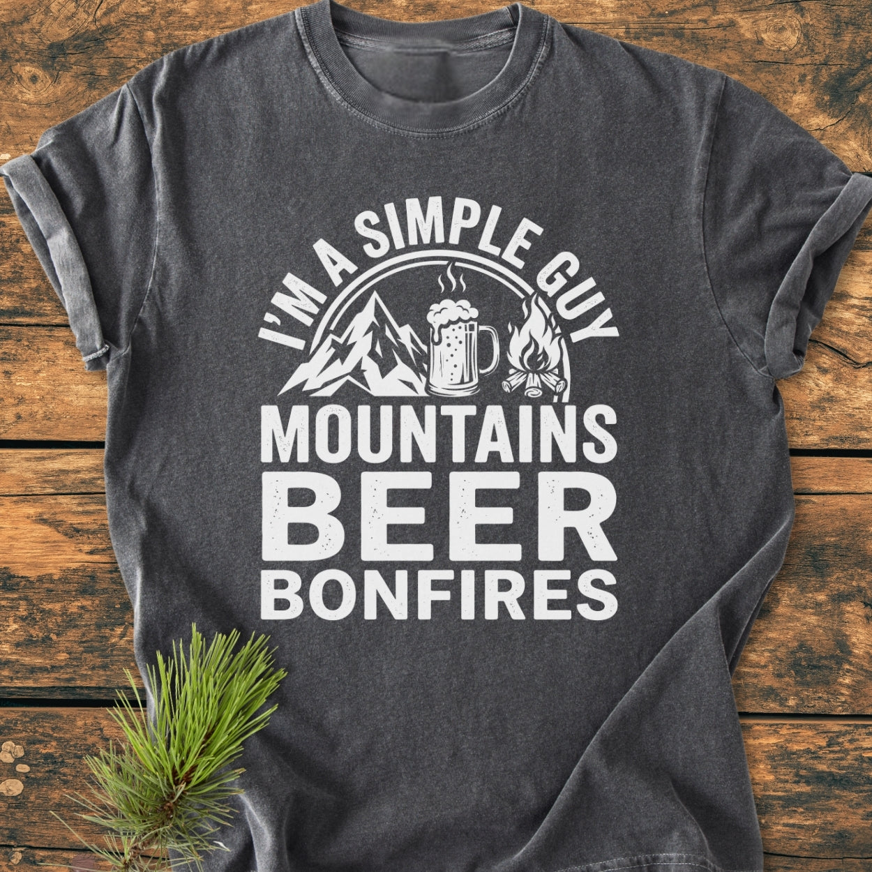 Mountains Beer Bonfires - Vintage Wash Tee: Charcoal-gray, short-sleeve T-shirt with white graphics of mountains, beer mug, and campfire, laid on wood surface with pine sprig, emphasizing outdoor adventure themes.