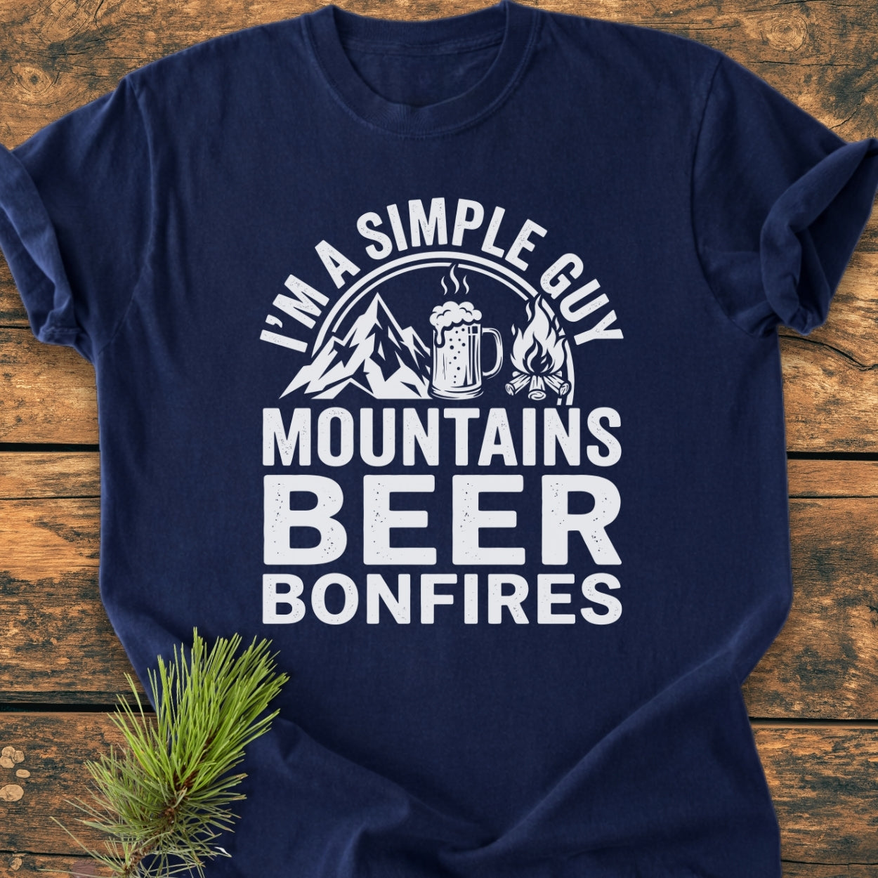 Mountains Beer Bonfires - Vintage Wash Tee featuring a white graphic of mountains, beer mug, and campfire on navy-blue fabric, laid on wood with pine sprig, highlighting its relaxed fit and durability.
