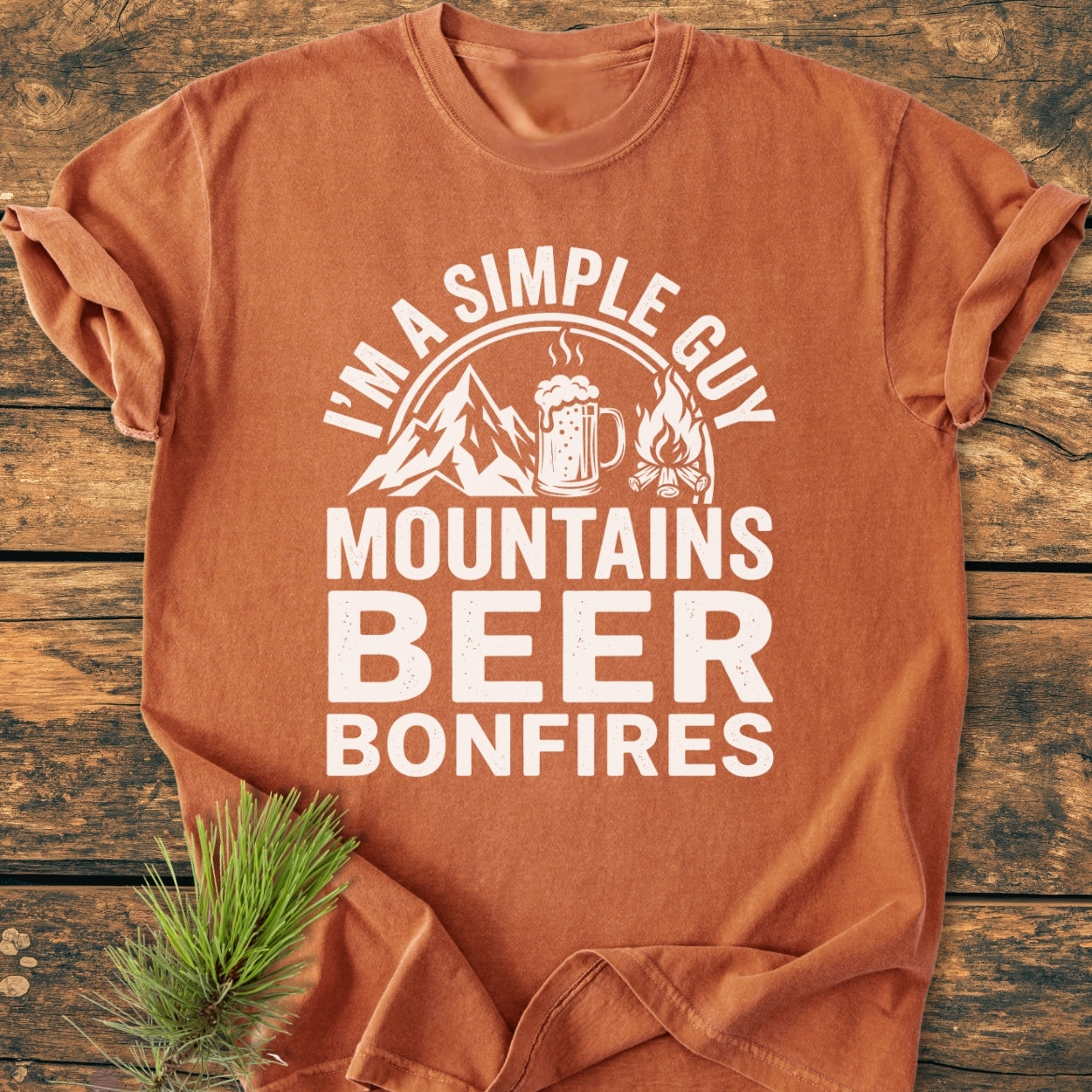 Mountains Beer Bonfires - Vintage Wash Tee; features a mountain, beer mug, bonfire graphic. Laid on wood with a pine sprig, showcasing its cozy, relaxed fit and durable stitching.