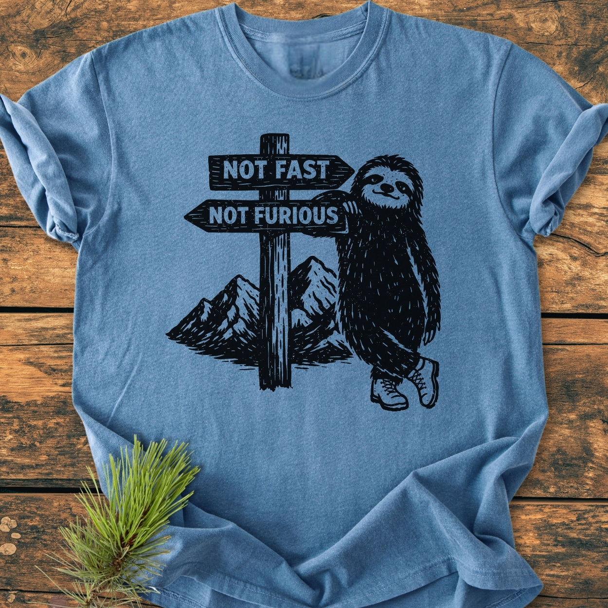 Not Fast Not Furious - Vintage Wash Tee features a cartoon sloth in hiking boots with a signpost, on a light-blue, soft-washed, garment-dyed T-shirt, laid on a wooden surface.