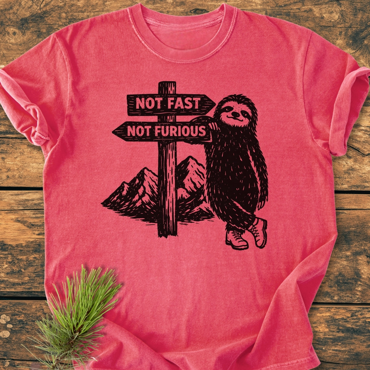 Not Fast Not Furious - Vintage Wash Tee featuring a sloth in hiking boots leaning against a signpost, laid on a rustic wooden surface with a pine sprig, showcasing its relaxed fit and durable stitching.