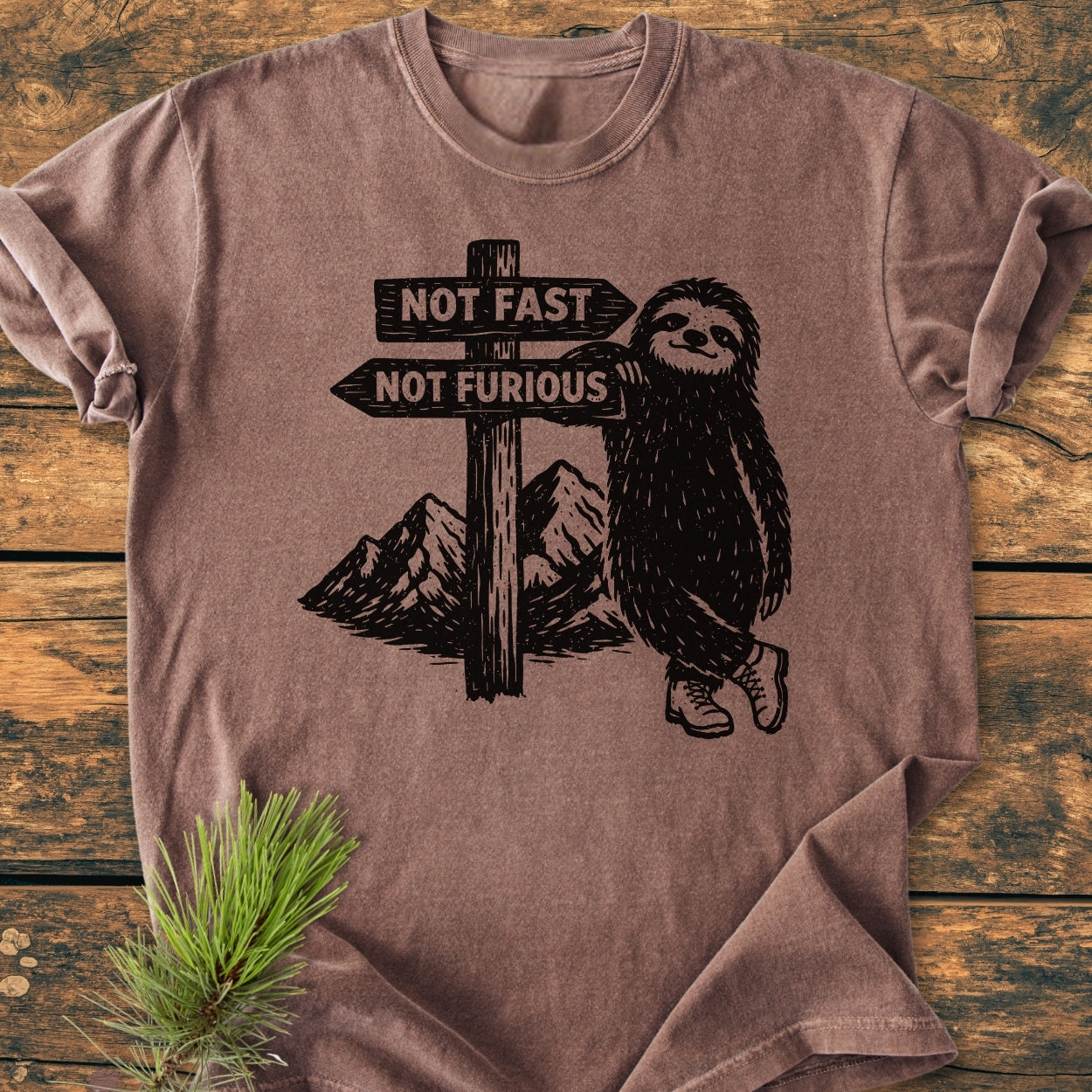 Not Fast Not Furious - Vintage Wash Tee featuring a sloth illustration with hiking boots and a signpost, laid on a wooden surface, complemented by a pine sprig for a rustic touch.