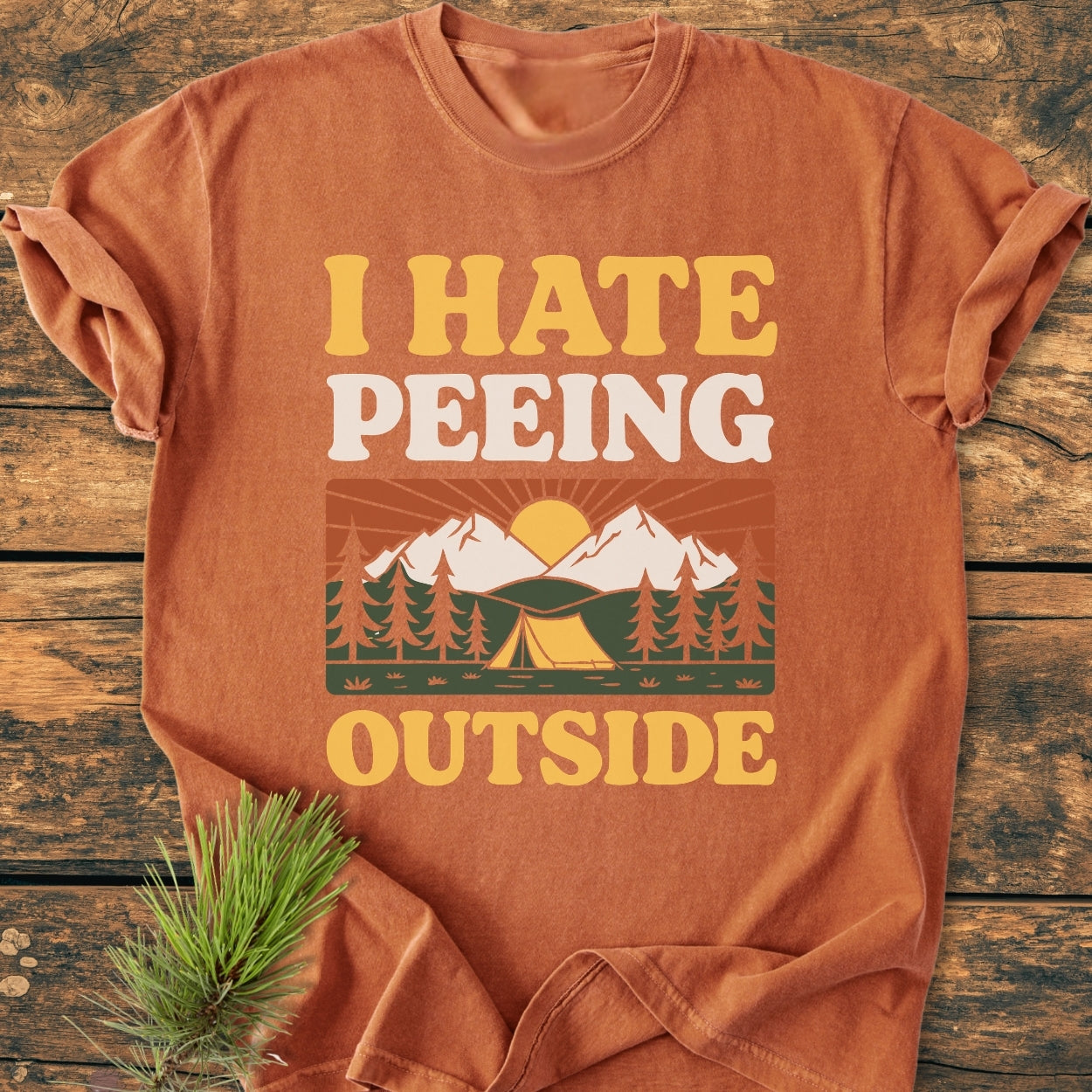 Folded I HATE Peeing Outside - Vintage Wash Tee in orange-rust, featuring a slogan with an outdoor scene of a tent, pine trees, and mountains. Placed on a wooden surface with a pine sprig beside.