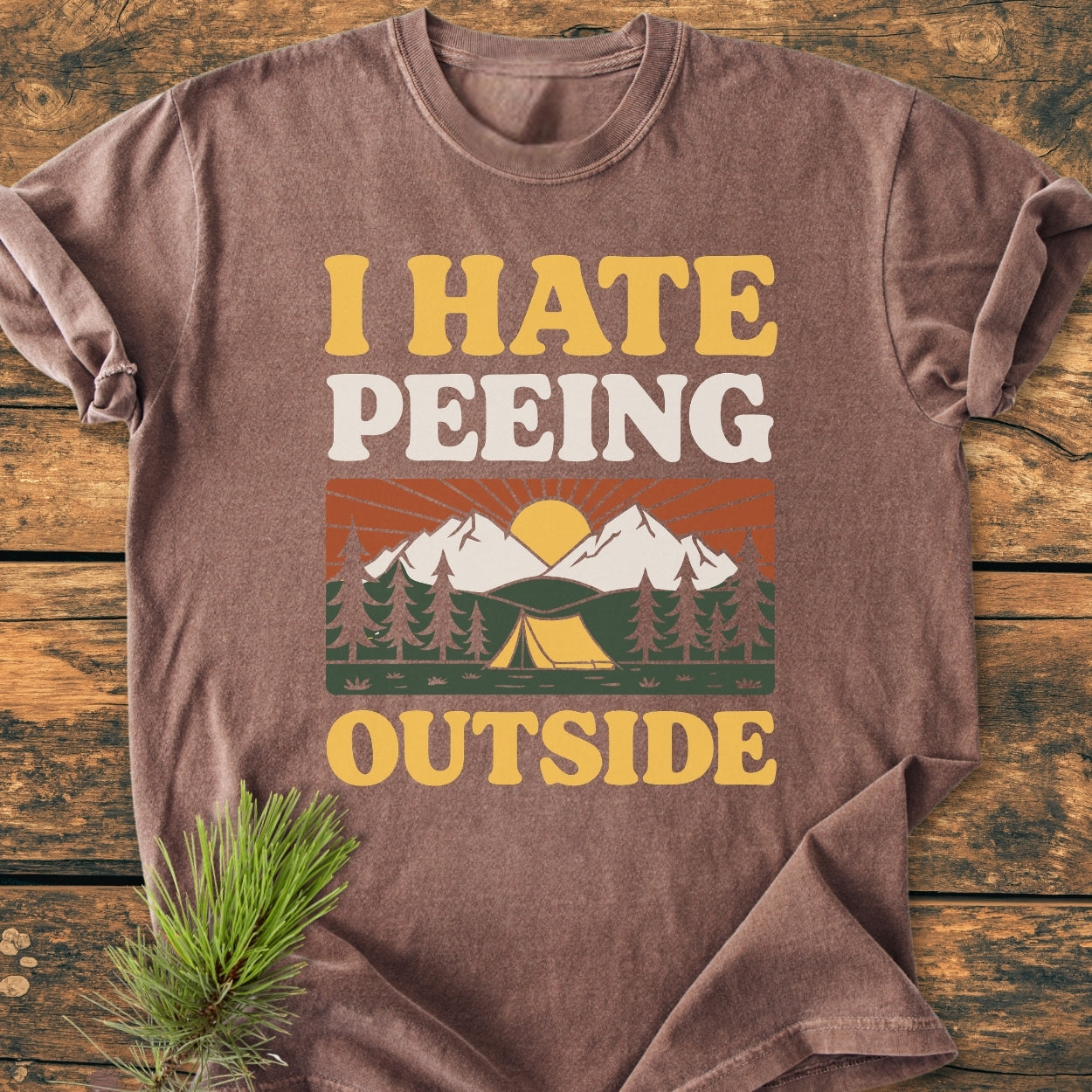 Brown I HATE Peeing Outside - Vintage Wash Tee with retro camping scene, laid flat on wood. Features bold text and graphics, made from soft-washed, garment-dyed 100% ring-spun cotton for relaxed comfort.