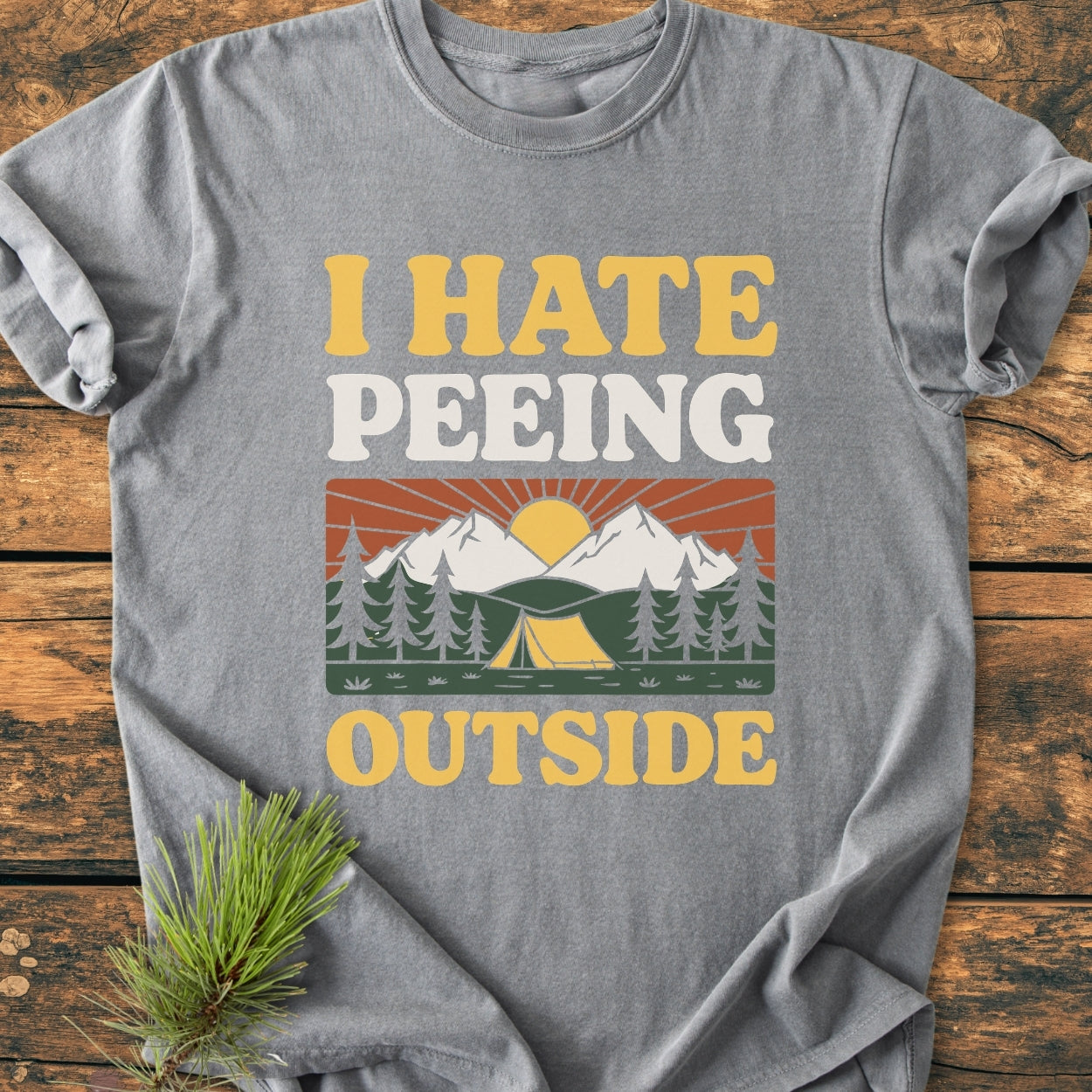 Heather-gray I HATE Peeing Outside - Vintage Wash Tee featuring a retro camping graphic with mountains, trees, and tent; laid flat on wood with a pine sprig, emphasizing its outdoor theme.