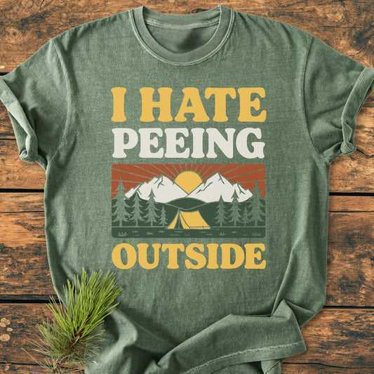 Forest-green I HATE Peeing Outside - Vintage Wash Tee features a retro camping scene with a yellow tent and pine trees, laid on wood with a pine sprig, highlighting its relaxed fit and soft fabric.