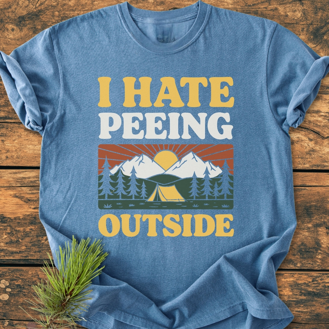 I HATE Peeing Outside - Vintage Wash Tee featuring a camping scene with pine trees, tent, mountains, and sun on a blue T-shirt, laid on wood, with a pine branch nearby.