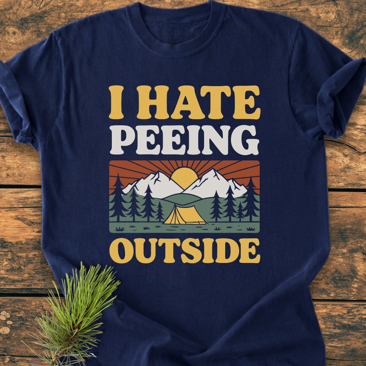 Navy-blue I HATE Peeing Outside tee features a retro camping scene with a yellow tent, pine forest, and mountains. Soft-washed, relaxed fit, pre-shrunk cotton, laid flat with pine sprig beside.