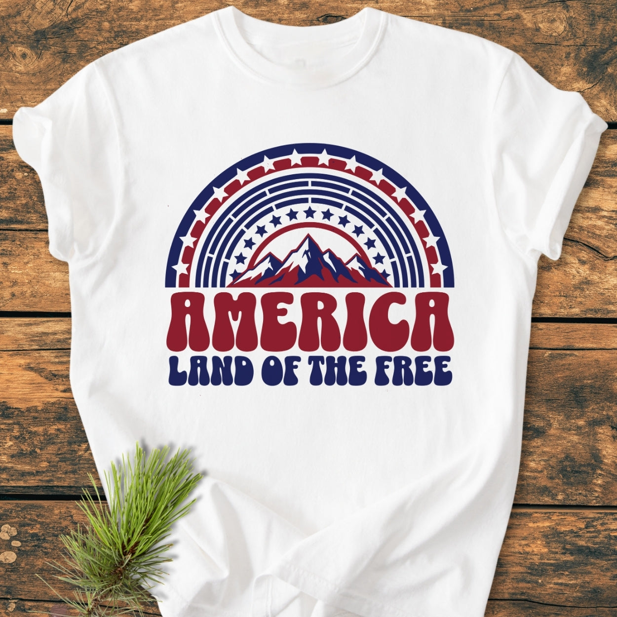 America Land Of The Free - Vintage Wash Tee; white T-shirt with retro red-white-blue stars and mountain graphic, laid on rustic wood, showcasing its soft, garment-dyed fabric and relaxed, durable construction.
