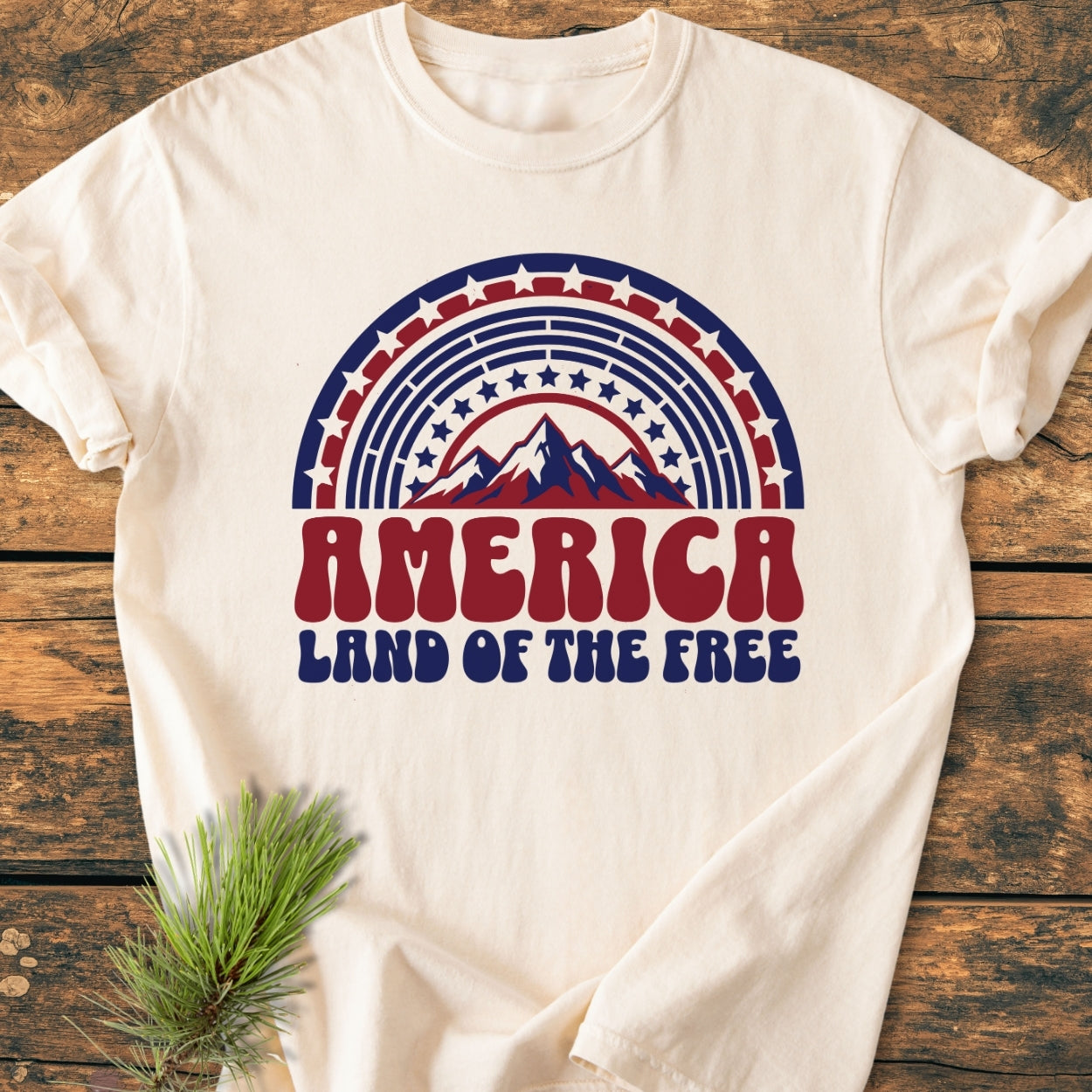 America Land Of The Free - Vintage Wash Tee displayed on a rustic wooden surface, featuring retro red and blue graphics with stars and a mountain motif, complemented by a small pine branch accent.