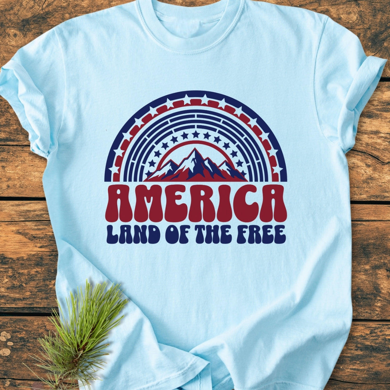 America Land Of The Free - Vintage Wash Tee features a retro-style graphic with a semicircular rainbow of stars and mountain silhouette, laid flat with a pine bough under one sleeve on wood.