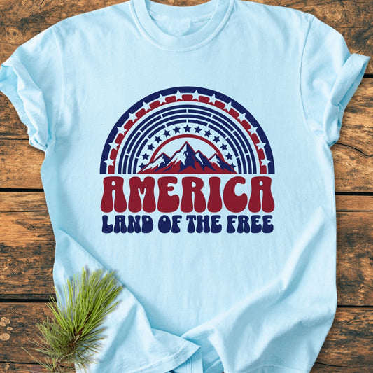 America Land Of The Free - Vintage Wash Tee features a retro-style graphic with a semicircular rainbow of stars and mountain silhouette, laid flat with a pine bough under one sleeve on wood.