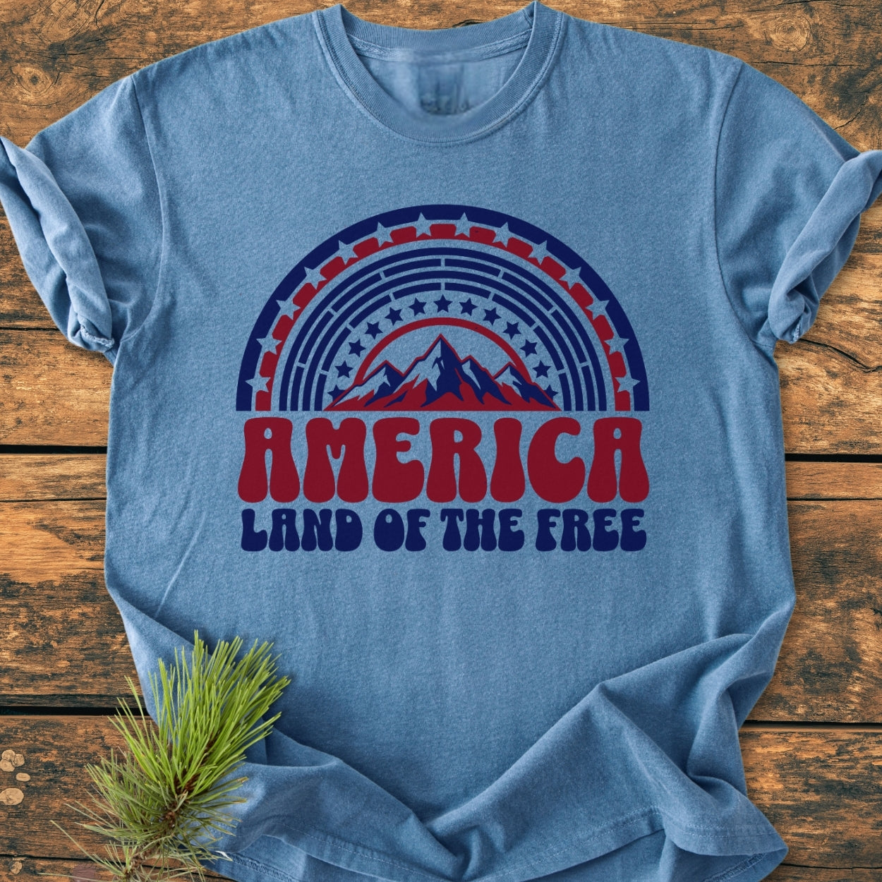 Heather-blue America Land Of The Free - Vintage Wash Tee featuring retro red and navy graphic with stars, stripes, and mountain silhouette, laid flat on wood with pine sprig beside.