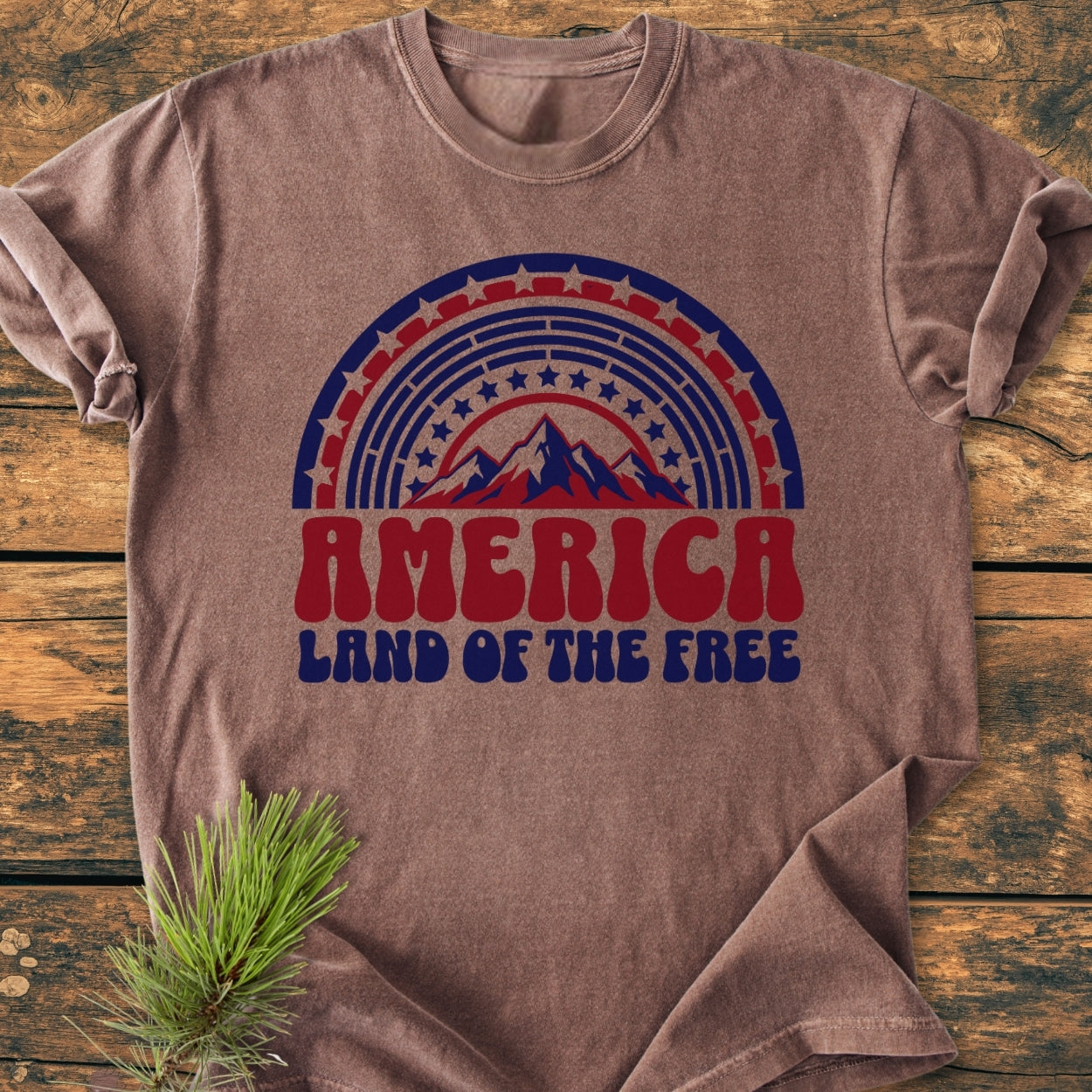 America Land Of The Free - Vintage Wash Tee featuring retro mountain peaks and patriotic stripes design on a soft, relaxed-fit, earth-tone T-shirt, laid flat with a pine sprig accent.
