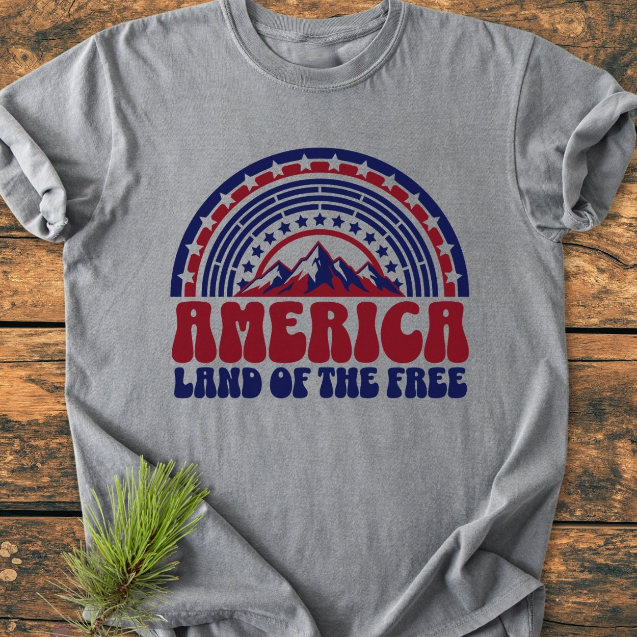America Land Of The Free - Vintage Wash Tee featuring a retro graphic with mountains and stars, laid on a rustic surface with a pine sprig, showcasing its relaxed, durable, ring-spun cotton design.