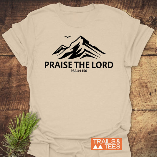 Praise The Lord, Psalm 150 T-Shirt featuring a black stylized mountain and bird graphic centered on light-cream fabric, laid flat with an orange-and-white TRAILS & TEES logo nearby.