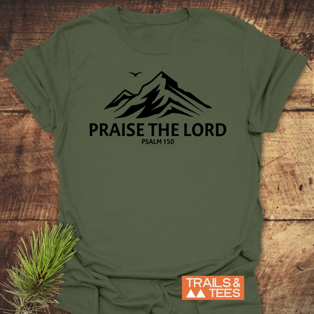 Olive-green Praise The Lord, Psalm 150 T-Shirt with mountain silhouette and bird, laid on wood. Accompanied by Trails & Tees logo and pine sprig, showcasing an outdoorsy feel.