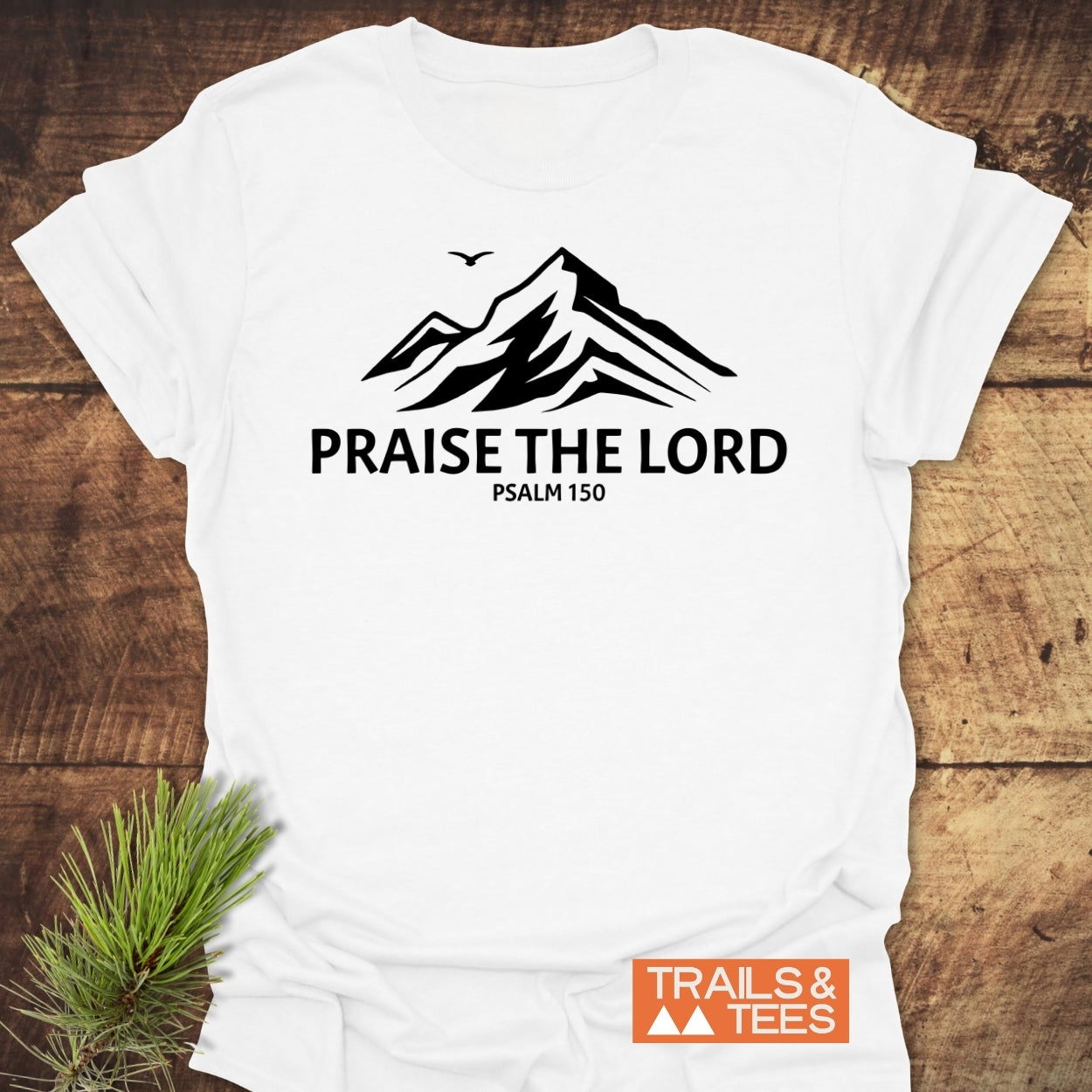 Praise The Lord, Psalm 150 T-Shirt featuring black mountain peaks and a bird graphic. Laid on wood, with a pine sprig, showcasing timeless design and comfort from Trails & Tees.