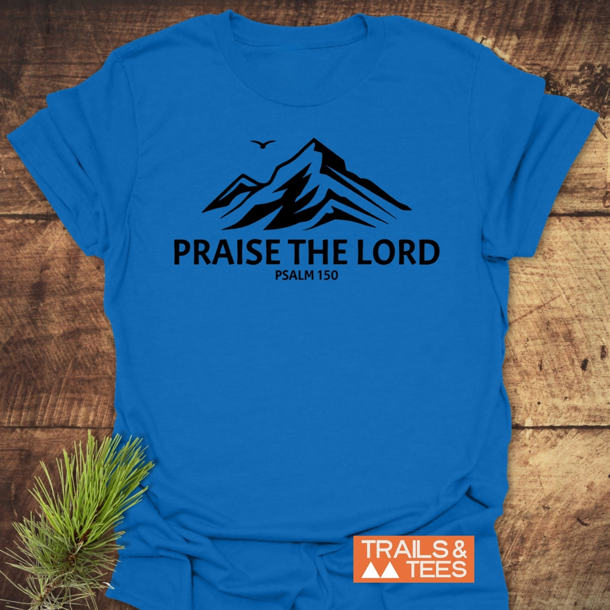 Praise The Lord, Psalm 150 T-Shirt featuring a mountain silhouette and bird design. Laid flat with a pine sprig, showcasing its trail-tested comfort and durable print from Trails & Tees.