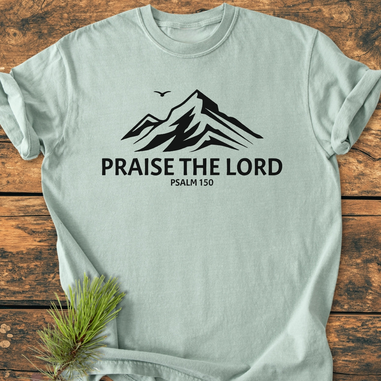 Praise The Lord, Psalm 150 - Vintage Wash Tee featuring a mountain graphic and bird silhouette, laid on wood with pine sprig, showcases a relaxed fit, durable stitching, and soft garment-dyed cotton.