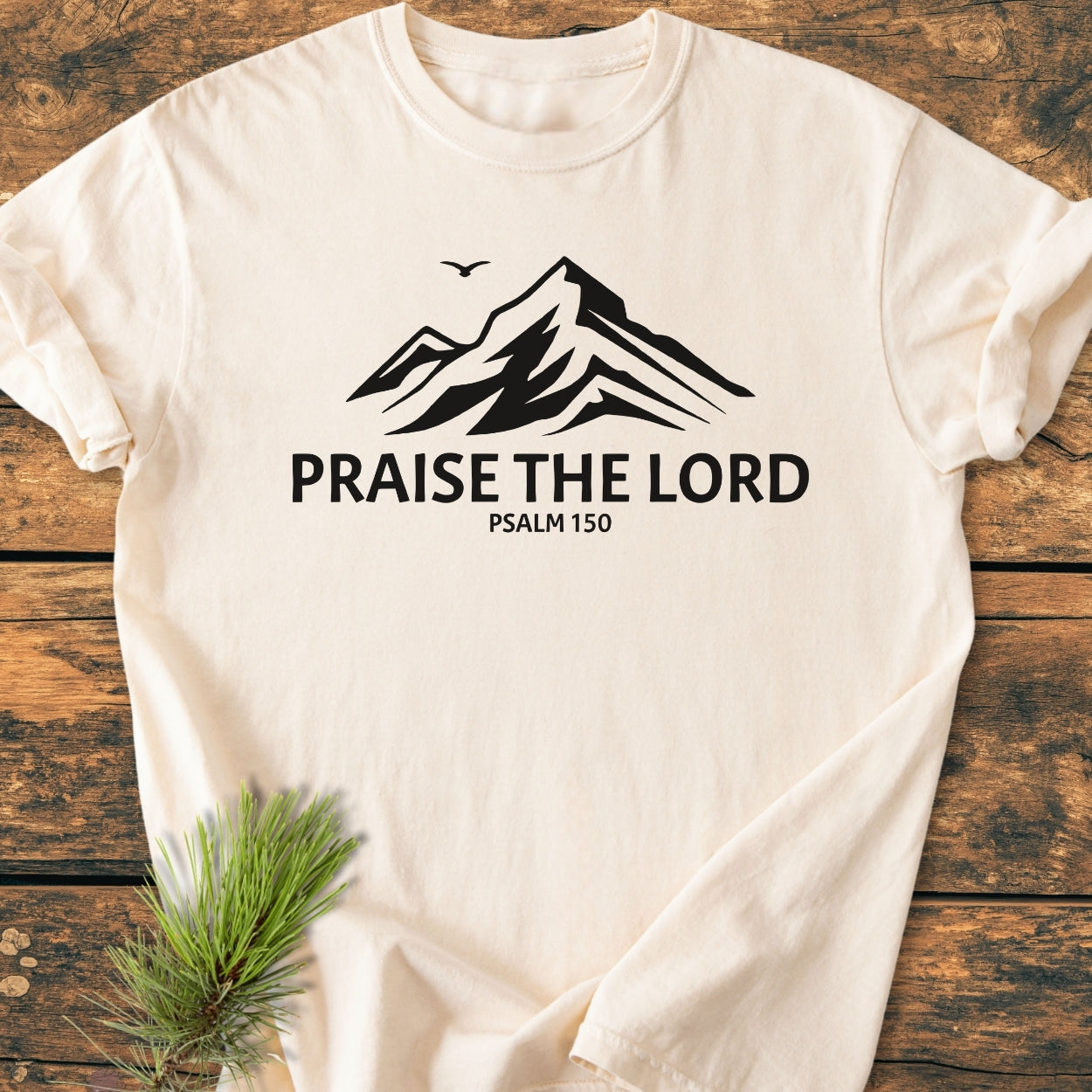 Praise The Lord, Psalm 150 - Vintage Wash Tee features a black mountain peak graphic with a bird silhouette and text on a cream T-shirt, laid on wood with a pine branch.
