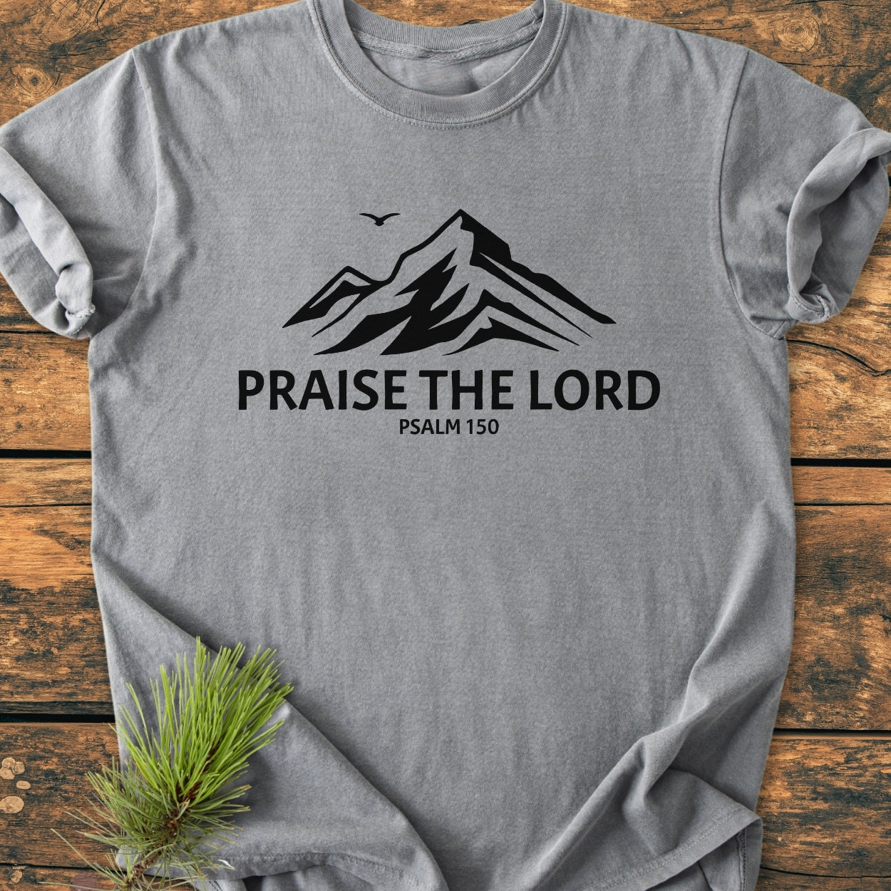 Praise The Lord, Psalm 150 - Vintage Wash Tee features a mountain silhouette with a bird and bold text, laid on wood. A pine sprig accents the soft, durable, relaxed-fit design.