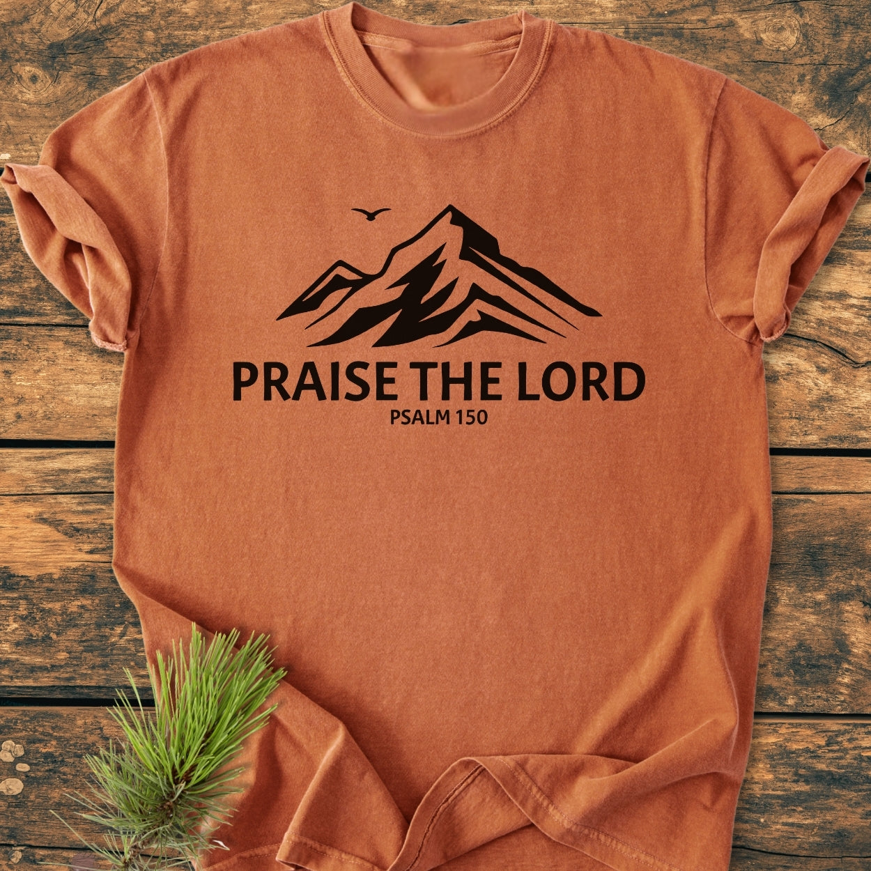Praise The Lord, Psalm 150 - Vintage Wash Tee features a stylized mountain graphic with a bird silhouette, and text, on an orange-rust T-shirt, laid on wood, accompanied by a pine sprig.