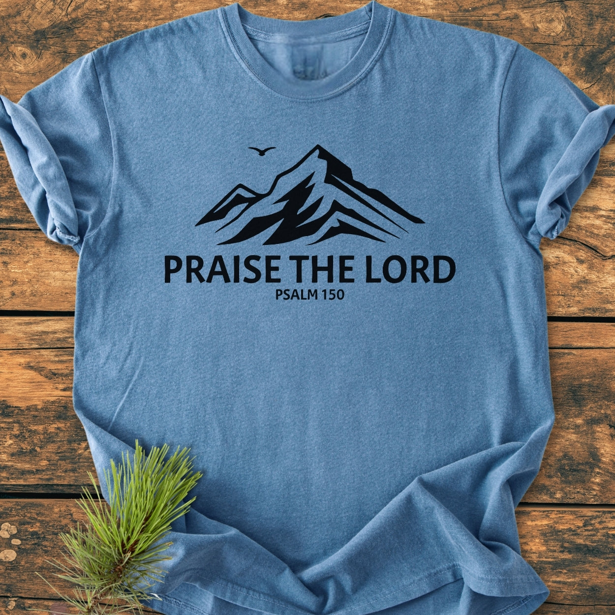 Praise The Lord, Psalm 150 - Vintage Wash Tee features a heather-blue hue with a mountain graphic and text, laid on wood with a pine sprig. Relaxed fit, soft, durable Comfort Colors garment.