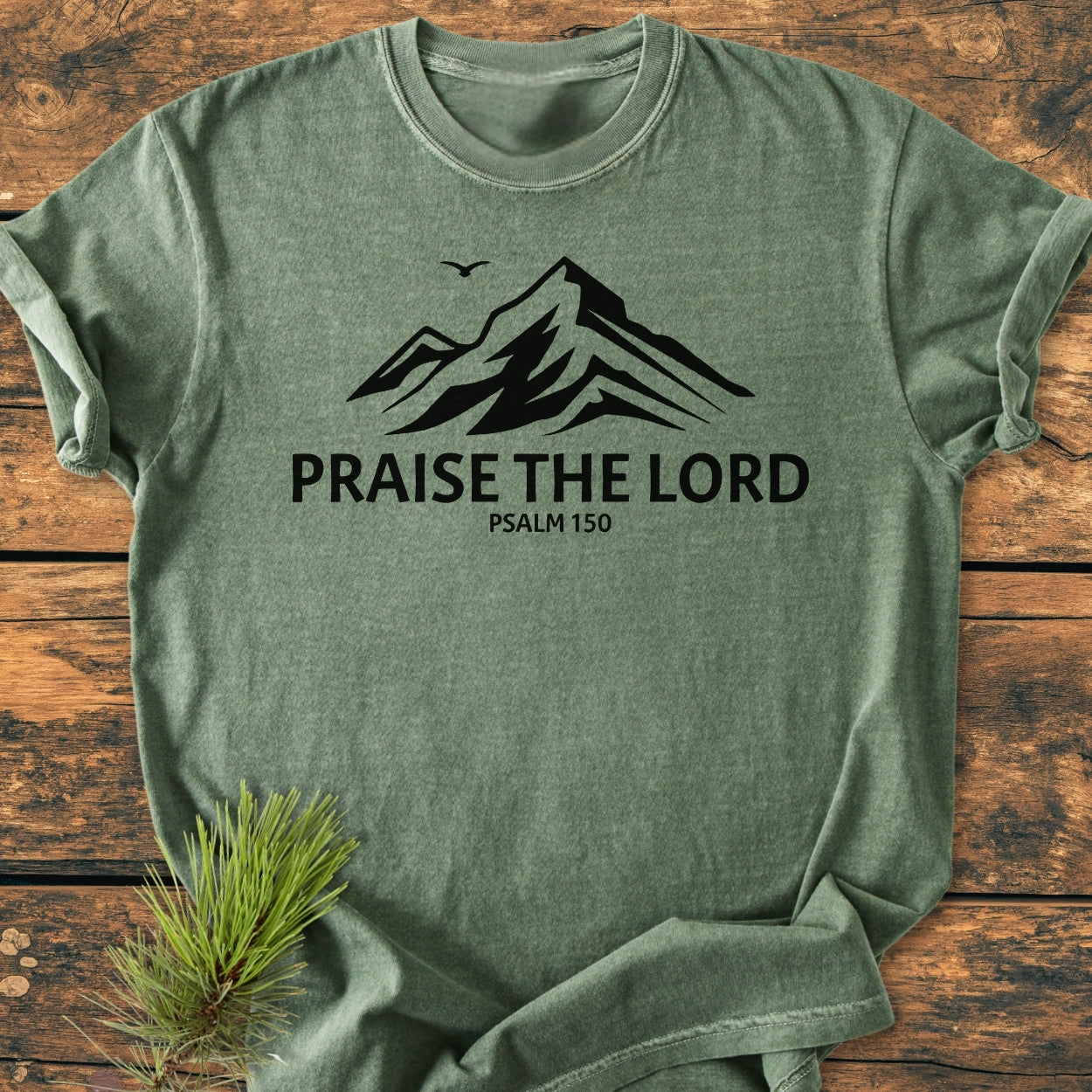 Praise The Lord, Psalm 150 - Vintage Wash Tee: Soft green crew-neck t-shirt with mountain silhouette and bird. PRAISE THE LORD and PSALM 150 printed below. Laid on wooden surface with pine sprig.