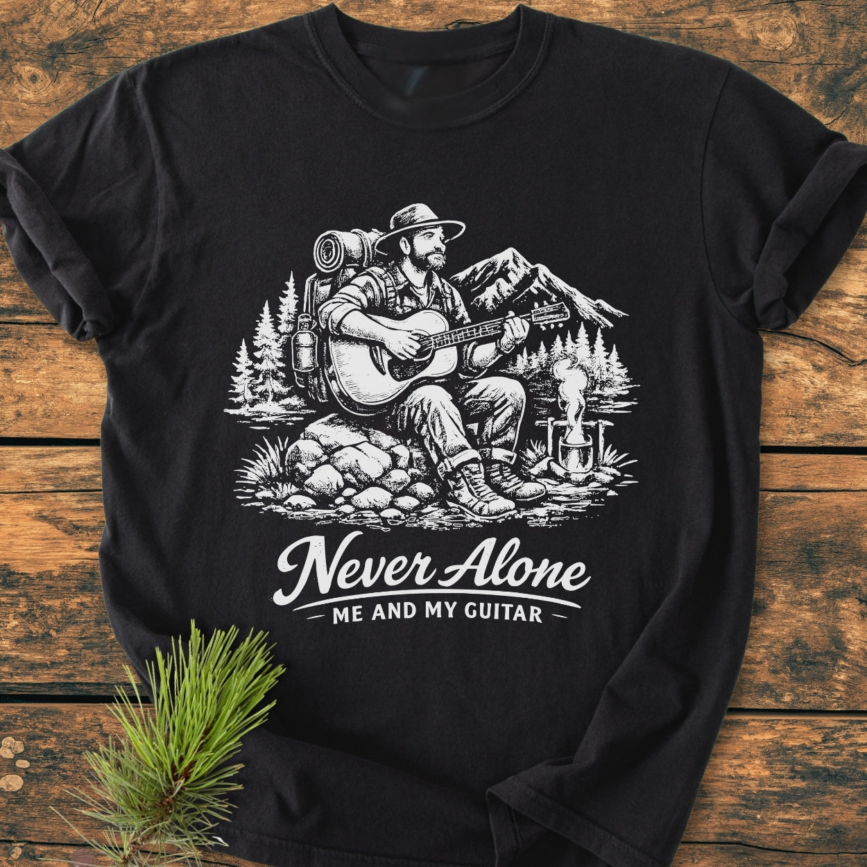 Never Alone. Me And My Guitar - Vintage Wash Tee features a bearded man by a campfire, playing guitar amid mountains and pines, printed on a soft, relaxed-fit, black T-shirt.