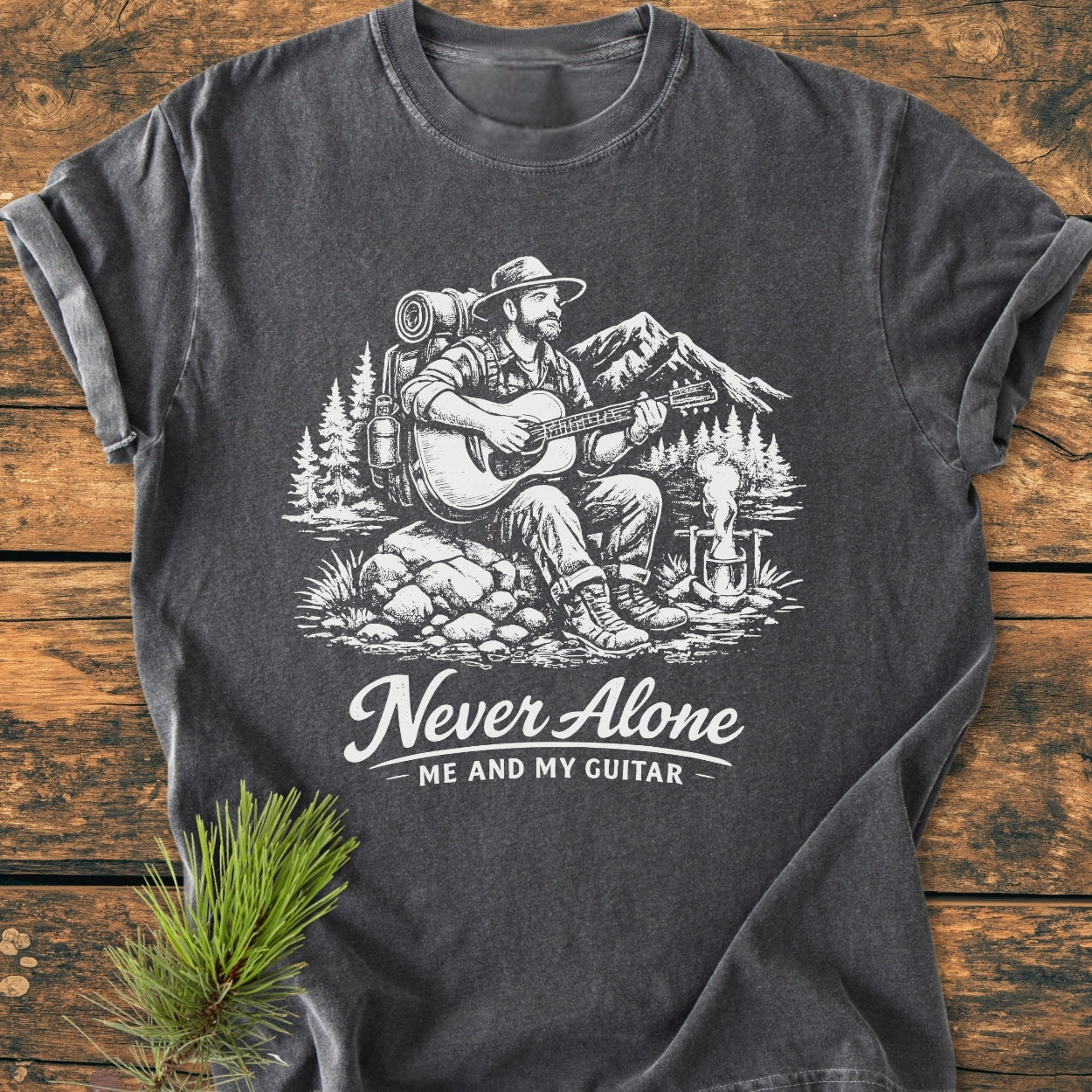 Never Alone. Me And My Guitar - Vintage Wash Tee features a hand-drawn scene of a traveler with guitar and campfire, set against trees, lake, and mountains, printed on a charcoal-gray T-shirt.