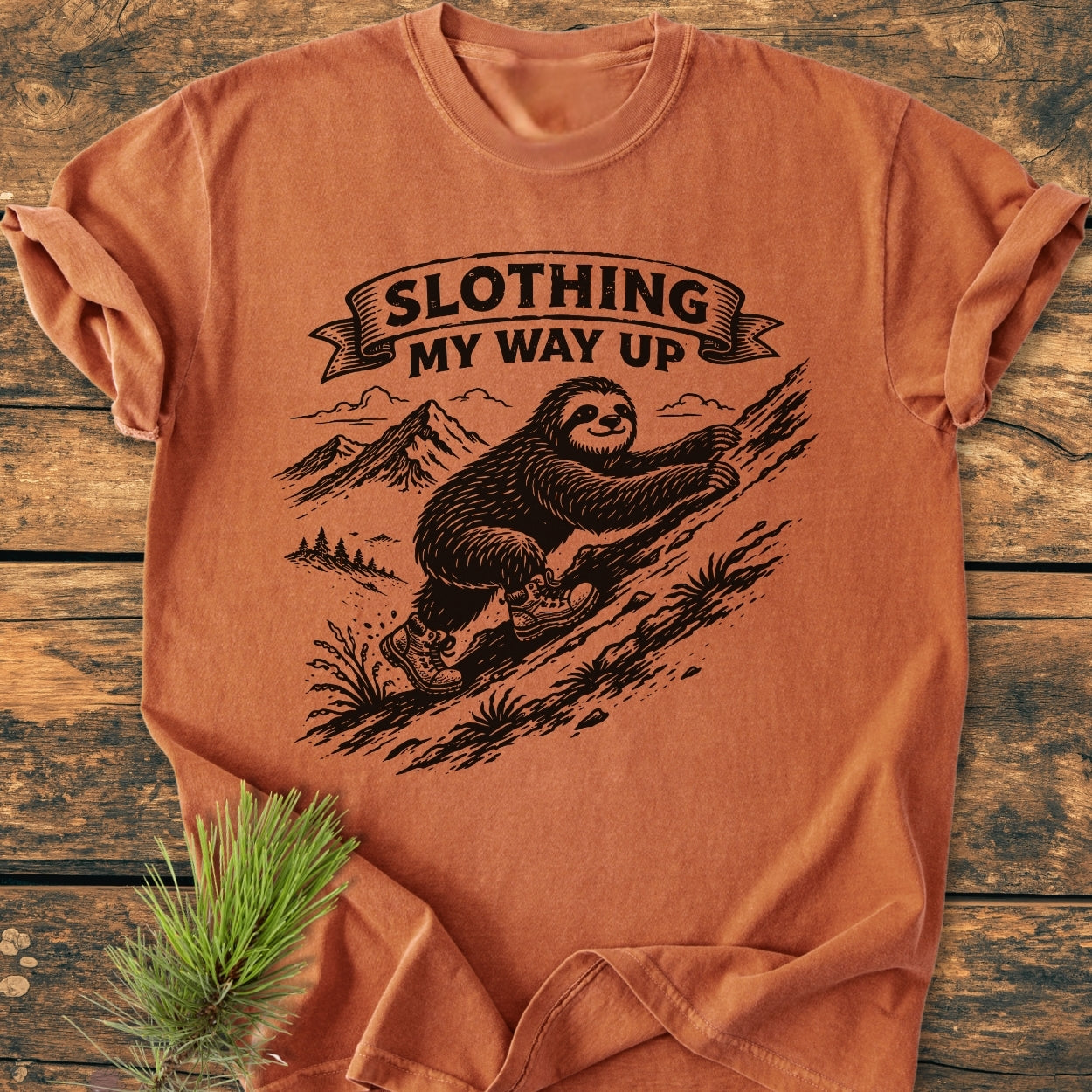 Slothing My Way Up - Vintage Wash Tee featuring a cartoon sloth in hiking boots on a hillside, laid-flat on a wooden surface with a pine sprig, showcasing its relaxed fit and soft-washed fabric.