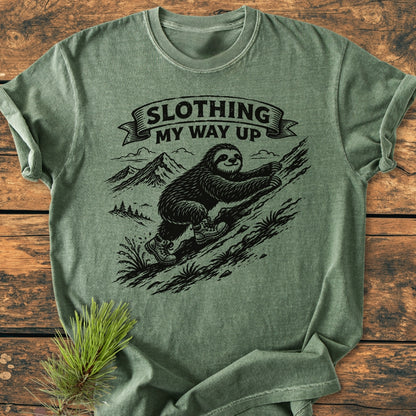 Slothing My Way Up - Vintage Wash Tee features a cartoon sloth hiking mountains, printed on a heather-green shirt, laid on wooden surface with pine sprig, highlighting its cozy, durable design.