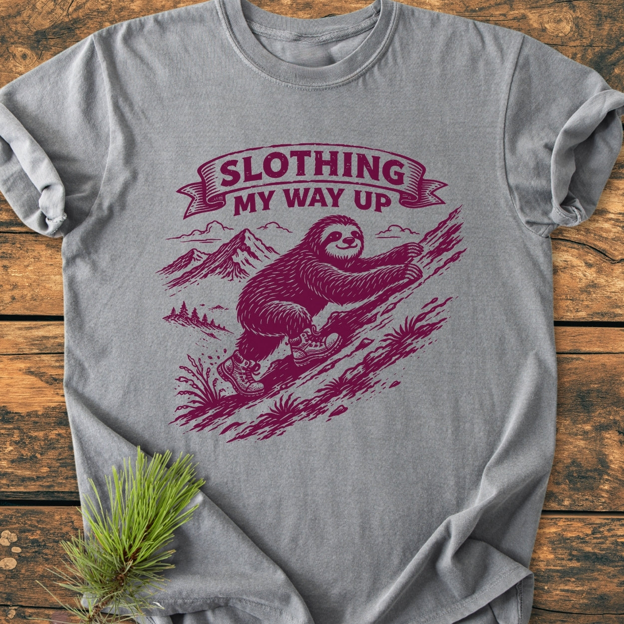 Slothing My Way Up - Vintage Wash Tee features a cartoon sloth in boots climbing a slope, printed on a heather-gray T-shirt, styled with a pine sprig accent on a wooden surface.