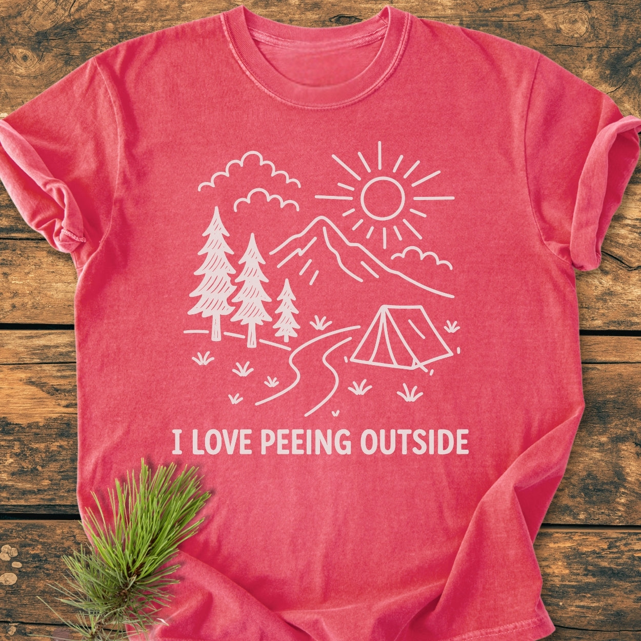 I Love Peeing Outside - Vintage Wash Tee featuring a line-drawn camping scene with sun, mountains, and tent. Displayed on a wooden surface with a pine branch beside it.