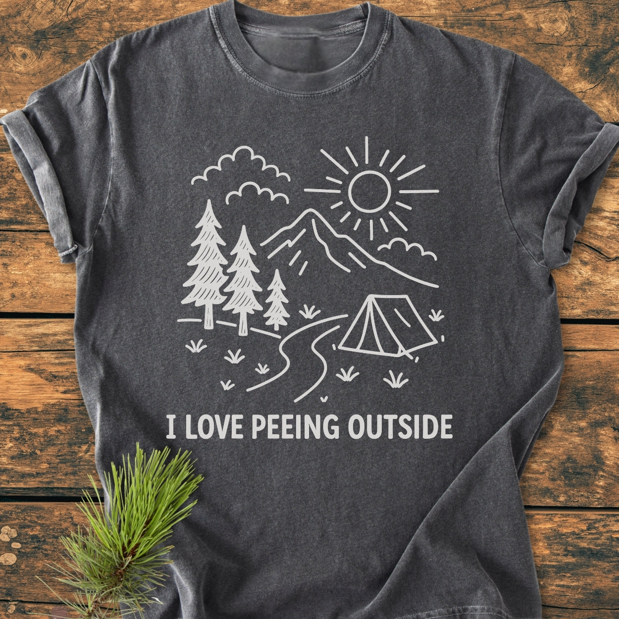 I Love Peeing Outside - Vintage Wash Tee showcases a line-art camping scene on a dark-grey shirt, emphasizing outdoor humor. Laid flat with a pine branch for a rustic touch.