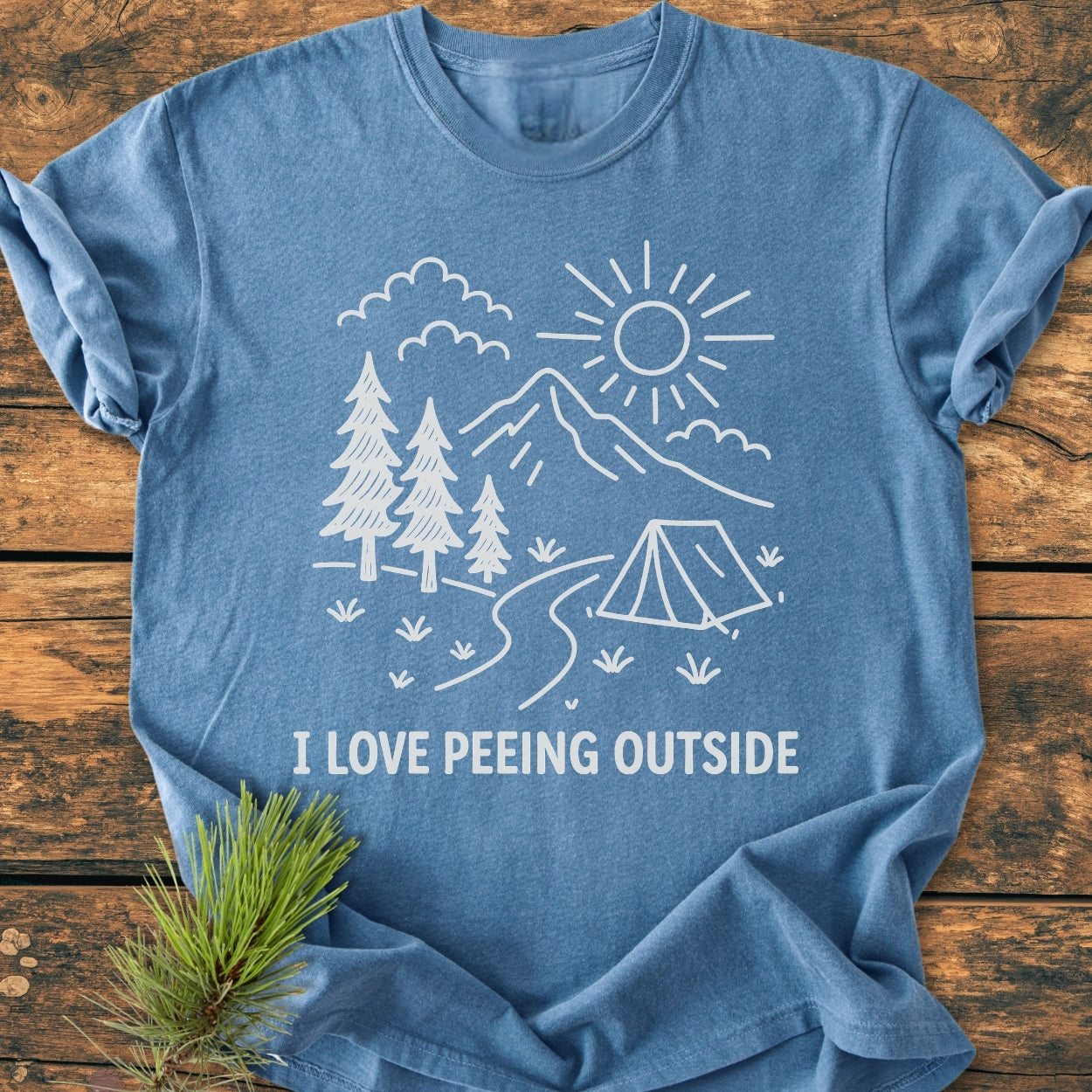 The I Love Peeing Outside - Vintage Wash Tee displays a line-drawn outdoor scene with pine trees and a tent, printed on a heather-blue T-shirt laid flat on a wooden surface.