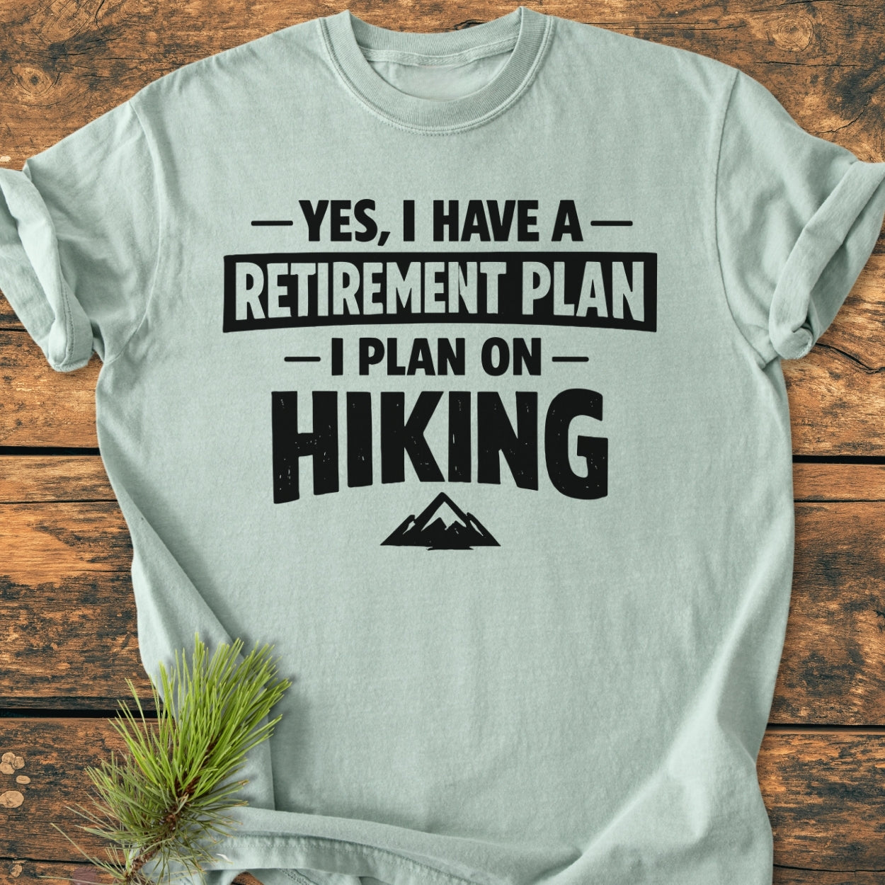 Retirement Plan Hiking - Vintage Wash Tee, featuring bold black lettering and a mountain silhouette, is laid on wood with a pine sprig, showcasing its soft, relaxed fit and durable, seamless design.